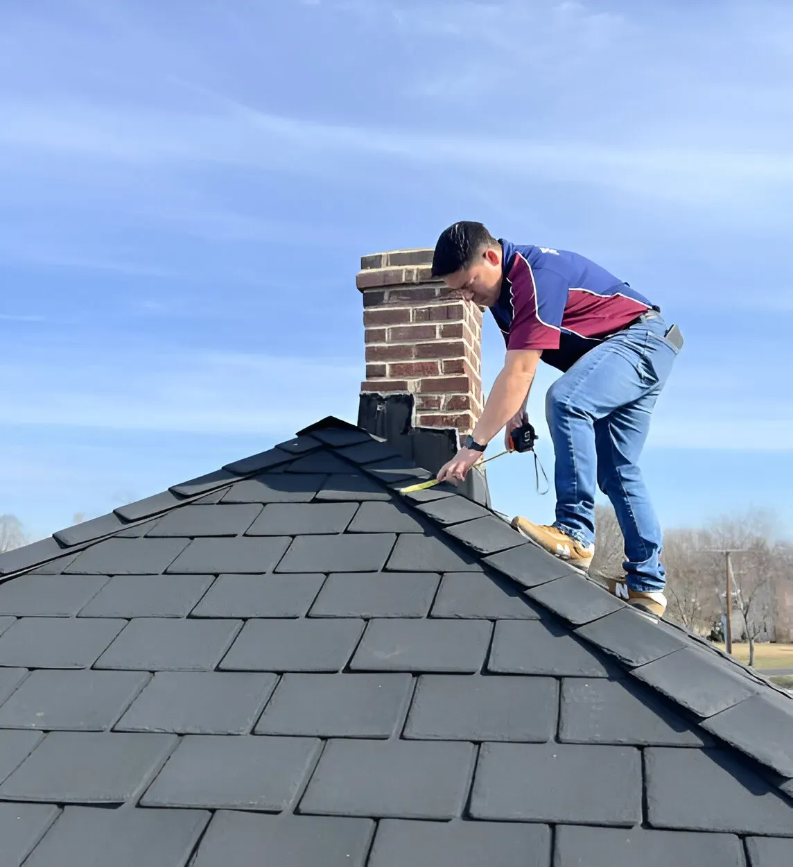 Worker applying finishing touches on a black shingle roof near a chimney.
