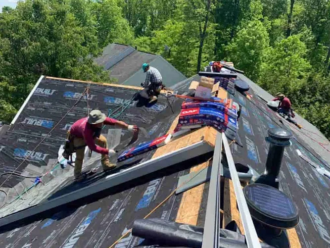 Workers installing roofing materials on a sloped residential roof surrounded by trees.